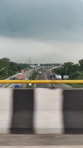 The video shows a view from a moving vehicle, looking over a highway. The sky is overcast with dark clouds. The highway is busy with traffic, including trucks and cars. There are trees and other vegetation on either side of the highway. The foreground of the video shows a blurry railing with black and white stripes. The camera is panning slightly to the right.