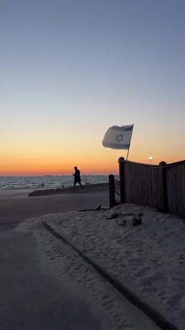 A static video captures a beach scene at sunset. The sky is a gradient of blue, orange, and yellow. On the left, a person walks along a paved path next to the ocean. The ocean is calm with small waves reflecting the sunset. On the right, a wooden fence stands next to a sandy area. An Israeli flag, blue and white with a Star of David, is attached to a pole on the fence, flapping gently in the breeze. A street lamp is visible behind the fence. Another person walks past the fence from right to left. The sand in the foreground shows a slight incline and some debris.