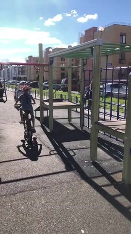 A child rides a bicycle on a paved playground while another child climbs on a jungle gym structure. The jungle gym has green monkey bars and blue netting. The sun is shining, casting shadows on the ground. Buildings and cars are visible in the background.