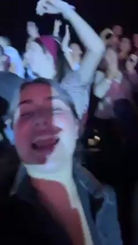 The video captures a live concert. Two young women hold up a phone to record themselves and the crowd in the background. The lighting shifts from white to dark to bright pink as the women cheer, sing along, and smile at the camera. Other concert-goers are visible in the background, standing and clapping along with the music. The video's perspective is from the viewpoint of the person holding the phone.