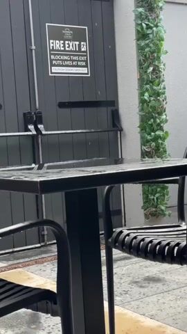 The video shows a black metal table with chairs in the foreground. Behind the table are black double doors with a "FIRE EXIT" sign on the left door. The sign reads "FIRE EXIT BLOCKING THIS EXIT PUTS LIVES AT RISK CAUTION: DOORS OPEN OUT." To the right of the doors is a tall pillar covered in green artificial ivy. The ground is wet, indicating it may be raining or has recently rained. The video is very dark and the scene appears to be outdoors.