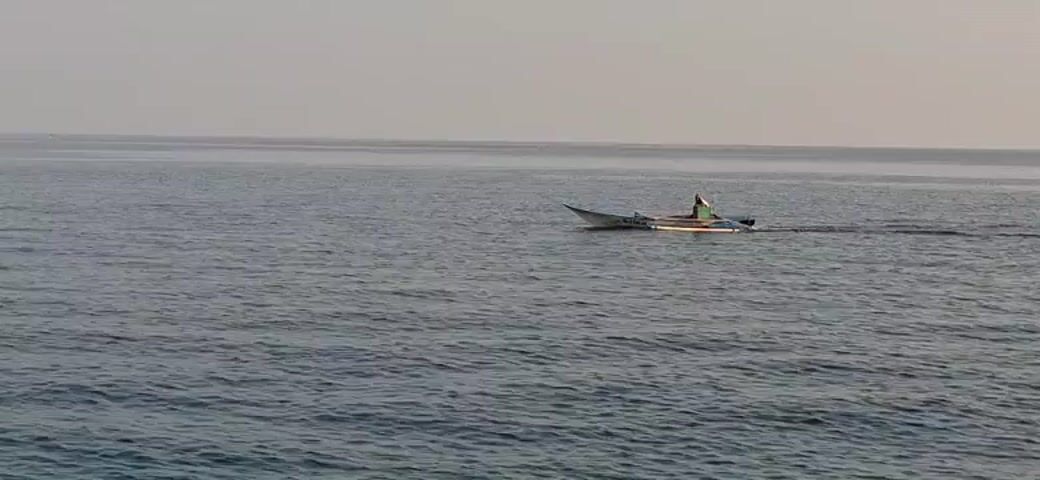 A small white boat with a blue stripe travels across calm water under a hazy sky. A single person wearing a green shirt and a hat is seated in the boat. The boat appears to be moving from left to right across the frame, with a visible wake trailing behind it. The horizon is visible in the distance, where the water meets the sky.