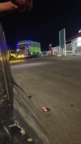A nighttime scene shows a truck with green tarped cargo and bright yellow headlights approaching the camera. Blue lights are visible on the truck's roof. A smaller yellow truck is visible behind it. The scene appears to be at a checkpoint or toll booth, with a white building and metal barriers in the background. A small, colorful object, possibly candy wrappers or a small piece of litter, is on the asphalt in the foreground. The perspective suggests the video is being recorded from behind a large, dark object, possibly a vehicle or barrier. The action is the truck moving forward.