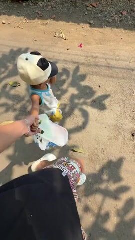 A child wearing a panda-themed hat, a light blue t-shirt, and yellow shorts is holding hands with a person. The child also appears to be holding a plastic bag. The person is wearing a black skirt and white sandals. They are walking on a dirt path with some debris on it, possibly in a rural or unpaved area. The sun is shining, casting shadows on the ground.