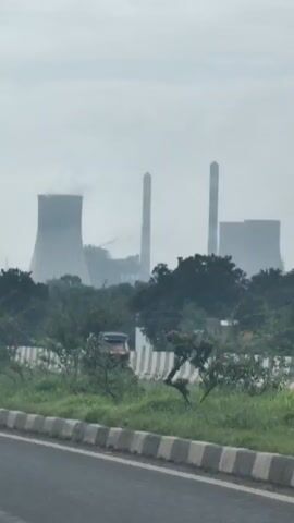 The video shows a view from a vehicle moving alongside a road. In the background, a large industrial complex with multiple tall structures, including cooling towers and chimneys, is visible. Smoke or steam is emanating from some of the structures, suggesting ongoing industrial activity, likely a power plant. The sky is overcast and hazy. In the foreground, there is a grassy area with trees and bushes, and a road barrier. On the right side of the road, some vehicles are visible, including what appears to be a truck. The overall impression is a landscape dominated by industrial infrastructure, viewed from a moving perspective.