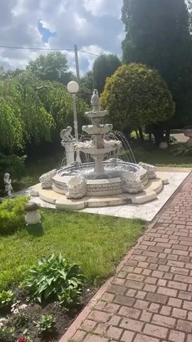 A tiered fountain with water flowing from each level is the central focus of the video. The fountain is ornate, with decorative elements and what appears to be carved lion heads around the base. The fountain is situated on a tiled platform in a garden setting. Lush green trees and foliage surround the fountain. The ground is covered with grass and a brick pathway is visible in the foreground. The sky is overcast with some clouds. There are no people in the video.
