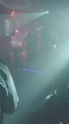 The video shows a group of people dancing on a small, elevated, illuminated platform in a crowded nightclub. The platform is lit up with a strip of bright purple lights.  The people on the platform are silhouetted against bright white stage lights.  The name "VALAR" is visible on a sign behind the platform. The environment is dark and hazy, and the crowd is made up of mostly young people. A person holding a camera is standing close to the dancefloor. Loud music plays in the background.