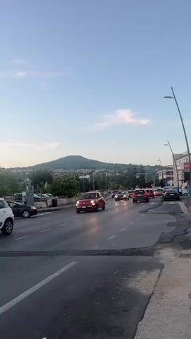This is a vertical, eye-level, medium shot of a street scene at dusk. The sky is a pale blue with a few wispy clouds. A large, tree-covered hill dominates the background. In the foreground, a multi-lane road is visible with several cars driving on it. The streetlights are on, casting a warm glow on the road and the vehicles. On the left side of the road, there are parked cars and some greenery. On the right side, there are buildings and a sidewalk. The overall impression is one of a peaceful evening commute in a suburban or semi-urban area.