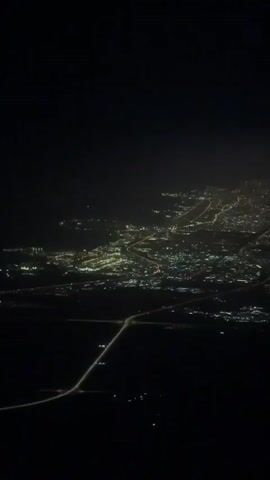 The video shows a night aerial view of a city. The city lights appear as bright dots and lines against the dark background. A highway with visible car lights runs diagonally across the lower part of the frame.