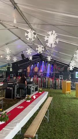 The video pans across a large tent, possibly for an event. In the background, a stage is set up with a band playing. There are lights illuminating the stage, and what appear to be speakers and sound equipment. In front of the stage, there are rows of skis standing upright, forming a decorative border. In the foreground, long tables are set up with white tablecloths and red runners. There are floral arrangements on the tables. People are seen working behind some of the equipment, and some band members are visible on stage. The tent ceiling is white, and large paper snowflake decorations hang from it. The ground appears to be covered in grass.