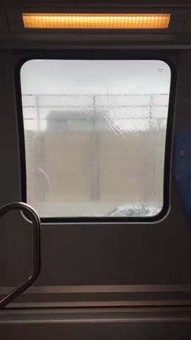 The video shows a view through a train window on a rainy day.  Rain streaks obscure the outside view, where concrete barriers and vegetation are visible in the blurred background. Inside the train, a handrail is visible to the left and a seat with blue patterned fabric is on the right. An overhead light illuminates the scene.