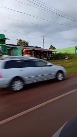 The video shows a roadside scene under an overcast sky. A road with a painted yellow line runs horizontally across the lower portion of the frame.  On the other side of the road is a grassy area with several buildings. The building closest to the road is a small structure with a dark roof and green tarpaulin on one side. It seems to be a small roadside shop with goods displayed. Further down the road is a green building, possibly another shop. There are people visible near these buildings, some walking, and others standing. A motorcycle speeds by on the road in front of the camera. Power lines stretch across the sky.