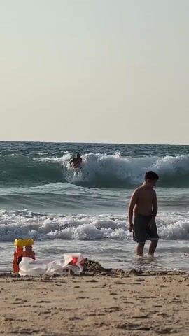 The video shows a beach scene with people in the water and on the sand. In the background, a surfer is riding a wave. In the middle ground, a man in swim trunks stands in the shallow water, looking out at the ocean. In the foreground, on the sandy beach, there are some discarded items, possibly plastic bags and toys. The sky is clear and the sun appears to be setting or rising, casting a warm light on the scene.
