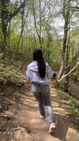 The video shows a group of people hiking on a dirt trail through a forest. The trees are green and leafy, and a stream runs alongside the path. The first part of the video shows the back view of a young woman with long dark hair wearing jeans and a white hoodie. Next, a young person with a black baseball cap is taking a selfie video. At one point in the video, the person with the cap points off to the side of the trail.