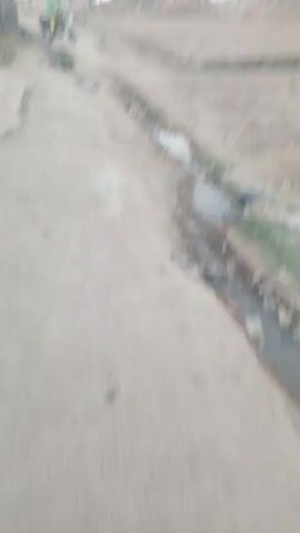 The video shows a person on a maroon motorcycle, parked on a dirt road next to a concrete structure or wall. The rider is wearing a green jacket and yellow pants. The road is cracked and shows signs of disrepair, with a shallow ditch running alongside it filled with dirty water and some trash. In the background, there are some distant buildings and trees under an overcast sky. The camera is held relatively steady, providing a clear but slightly distant view of the motorcycle and the surrounding environment.