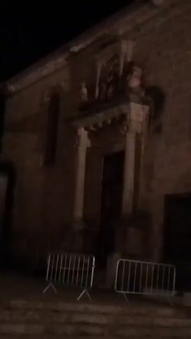 This video shows a building with a stone facade and an ornate entrance at night. There are two metal barricades in the foreground. A young man with dark hair and a black and white t-shirt walks past the building, looking up towards the entrance. The scene is dimly lit.