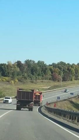 Two dump trucks are driving on a highway under a clear blue sky. The truck in the foreground is red and appears to be carrying a load of green material, possibly grass or foliage. The truck behind it is also red and is carrying a load of yellow material, possibly grain or sand. Both trucks are in the right lane of a multi-lane highway. Trees with autumn colors line the side of the road in the background. A white car is visible in the left lane in front of the dump trucks. There are traffic signs and a guardrail along the side of the highway. The video appears to be shot from inside another vehicle.
