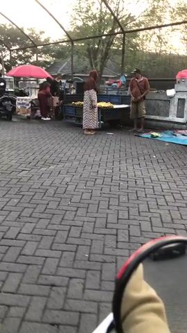 The video captures a lively street scene, likely in Boro, East Java, during what appears to be the late afternoon or early evening, given the warm, soft light filtering through the trees. The setting is an outdoor market or roadside area paved with interlocking grey bricks. Several food stalls are visible, with vendors attending to customers.

The central activity revolves around commerce and movement. People are gathered around the stalls, some examining goods, others interacting with vendors. Motorcycles, a common mode of transport in the region, are present in abundance, with some parked and others being ridden. One prominent stall features a bright red umbrella providing shade. Another stall has a display of what appears to be fresh produce, possibly bananas, arranged on a cart.

Individuals are dressed casually, with several women wearing headscarves, indicating a predominantly Muslim population. The general atmosphere seems bustling and active, suggesting a typical evening market or street food hub. The presence of various food carts and the flow of people and vehicles create a dynamic and vibrant picture of local life.

There is some visible text on a sign on the side of one of the motorcycles, but it is not legible enough to be deciphered. The overall mood conveyed is one of everyday activity and community engagement in an outdoor commercial space.