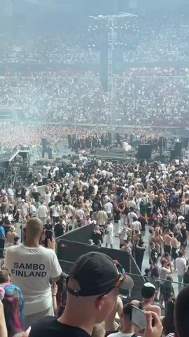 This media file captures a wide-angle view of a large outdoor concert or festival. A massive crowd of people fills the space in front of a stage, with many more spectators visible in the stadium seating surrounding the performance area. The atmosphere appears energetic and lively, with many individuals raising their hands and dancing.

The stage itself is a complex structure with scaffolding, lighting rigs, and what appears to be a drum set. Performers are visible on the stage, but they are small in the frame due to the distance. Smoke or haze is present in the air, particularly around the stage area, creating a dramatic effect.

In the foreground, the backs of several attendees are visible, with one person wearing a white t-shirt that clearly displays the text "SAMBO FINLAND". Another individual in the foreground is wearing a black baseball cap with colorful stripes. The overall scene suggests a summer event, as many people are dressed in light clothing. The lighting indicates it is daytime, likely late afternoon or early evening.

The location cue "FORTUNA DUSSELDORF" is visible in the upper background, indicating the event is taking place at the stadium of the German football club Fortuna Düsseldorf in Düsseldorf, Germany. The sheer number of people and the scale of the production point to a major event.