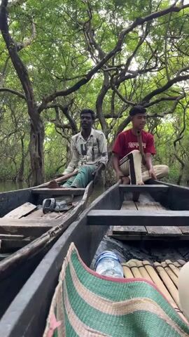 Two men are in separate boats, side by side, in a narrow waterway surrounded by dense forest. The man in the left boat, wearing a light-colored, patterned shirt and a green and blue striped lungi, is holding a long wooden oar. The man in the right boat, wearing a red t-shirt with black and white stripes on the sleeves and beige pants, is sitting cross-legged and holding a bamboo pipe to his mouth. Both men appear to be looking forward. The boats contain various items, including a water bottle, what appears to be a woven mat, and other unseen objects. The water is calm, and the trees overhead form a thick canopy, casting shadows. The atmosphere suggests a natural, perhaps rural, setting.
