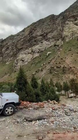This is a video clip of a mountainous landscape with scattered trees and a small structure. The scene appears to be in Gemur, India. The terrain is rugged, with a large, rocky mountain dominating the background. Green vegetation, including coniferous trees and low-lying bushes, is present on the slopes and in the foreground. A small, light-colored building with a red roof is visible amidst the trees. The sky is overcast, suggesting a cloudy or potentially rainy day. The overall atmosphere is one of natural grandeur and isolation. No people are visible in the clip. The video appears to be captured from a moving vehicle, as evidenced by the slight motion blur and changing perspective. The time of day seems to be daytime, but the cloud cover makes it difficult to determine the exact time. The location cues suggest a high-altitude, possibly remote area.