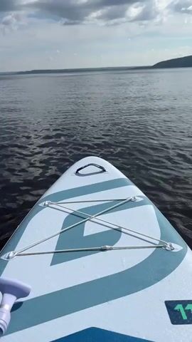 The video shows the bow of a paddleboard on a lake. The paddleboard is white with blue accents and has bungee cords stretched across the front. A purple paddle handle is visible on the left side of the frame. The water is dark and rippled. In the distance, a hazy shoreline can be seen under a cloudy sky.