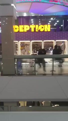 The video shows an indoor scene of a shopping mall or entertainment center. In the foreground, there is a glass barrier or railing with logos "MEGAPLANET" printed repeatedly. Behind the glass, there's a counter area with several people, potentially staff or customers. The counter is decorated in a purple and white color scheme. Above it, a large sign reads "CEPTION" in yellow, with what appears to be a sun or star graphic as the "O." Overhead, there is a sign in a Cyrillic language, possibly Russian, with the words "ДОБРО ПОЖАЛОВАТЬ" which translates to "WELCOME." Further back, there is a more elaborate structure with neon lights and another sign in Cyrillic, reading "ПАРК". The general atmosphere suggests an entertainment or game area. The scene is well-lit with overhead lighting. People are standing or seated behind the counter, and some appear to be looking at their phones. The video has a static viewpoint with no camera movement.