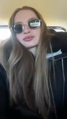 A young woman with long, straight brown hair is shown in a medium shot from the chest up. She wears square sunglasses with a gold-colored frame and a black zip-up jacket with white trim. Her lips are made up in a shiny mauve color, and her nails are painted red. She sits in what appears to be the passenger seat of a car, with the car's interior visible behind her. The light is bright, suggesting it's daytime. She makes a hand gesture, holding her hand up to her forehead with her fingers spread apart. Musical audio is present.