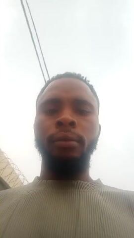 A person with a beard and short, dark hair looks into the camera. The person is wearing a light-colored ribbed shirt. The background includes a light, overcast sky with electrical wires, a wall with barbed wire, and an empty lot. The person raises their right hand to their forehead in what appears to be a salute.