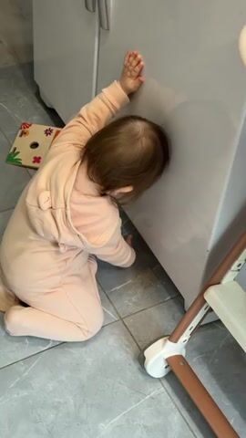A young child in a peach-colored hooded onesie is kneeling on a tiled floor, leaning against a white cabinet. The child's head is pressed against the cabinet door and their right hand is flat against the surface. A wooden toy is on the floor near the child. The leg of a highchair is visible to the right of the child.