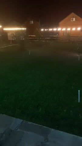 The video shows a dark outdoor scene with a grassy area and several buildings in the background illuminated by string lights. There are sprinklers visible in the grass. A paved path is in the foreground. The camera appears to be moving slightly.