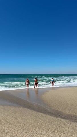 The video shows three women standing on a beach on a sunny day. All three are wearing swimsuits and facing the ocean. The woman in the center is standing with her arms outstretched. The sea has small waves. The beach is sandy. The sky is blue.
