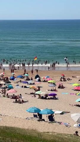 This is a high-angle, vertical, long shot of a crowded beach on a sunny day. The ocean is a vibrant blue-green with moderate waves and surfers. The beach is filled with people sunbathing, swimming, and relaxing under colorful umbrellas. There are several umbrellas scattered across the sand, some providing shade for groups of people. In the water, many people are swimming and wading. Lifeguard flags are visible on the beach, indicating a designated swimming area. The sand is light brown, and there are a few rocks visible near the bottom of the frame. The overall impression is a busy and lively beach scene on a hot summer day.