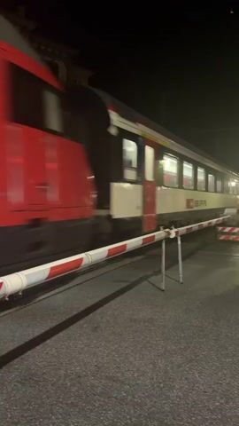 A nighttime video captures a train passing through a level crossing. The train is white with red and yellow stripes along the top, and it has several lit windows. The windows reveal red seats inside. The train has SBB CFF FFS branding visible. Red and white striped barriers are lowered across the road, indicating that the crossing is active. The road surface appears to be asphalt. The lighting suggests it is dark, with artificial lights illuminating the scene.