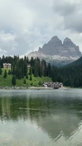 This media file captures a serene landscape featuring a calm lake in the foreground, reflecting the surrounding scenery. In the midground, a picturesque building with a white facade and brown roof sits on the shore, nestled amongst a dense forest of evergreen trees that ascend a lush green hillside. Beyond the trees, a majestic, jagged mountain range dominates the background, its peaks partially obscured by a cloudy sky. Several other buildings, smaller than the main one, are scattered on the hillside amongst the trees. 

The time of day appears to be daytime, with soft, diffused light suggesting overcast conditions. The weather is likely cool or mild, given the evergreen trees and the cloudy sky. The presence of a small boat on the lake and cars parked along the shore suggests human activity and a populated area, likely a tourist destination. The overall mood is peaceful and tranquil, showcasing the natural beauty of the mountainous region. The location is identified as Auronzo di Cadore, Italy, a region known for its stunning Dolomite mountain landscapes and lakes. There is no visible text in the media file.