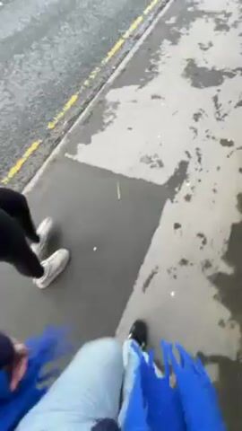 Two people are walking on a wet sidewalk next to a road. One person is wearing black leggings and light-colored sneakers, and the other is wearing light blue jeans and black and white sneakers. The road has cars driving on it, and there are houses and trees on either side of the road. The sky is cloudy.