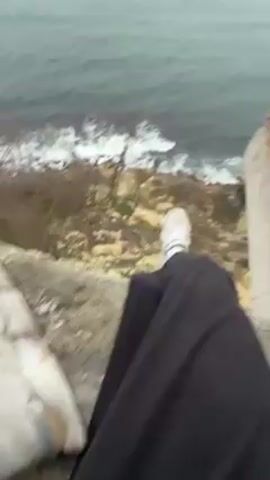 The video shows a first-person perspective of a person sitting on the edge of a crumbling building. The person is wearing black pants, white socks, and white sneakers. Below the building is a cliffside, and the ocean is crashing against the shore. The building appears to be abandoned and in a state of disrepair, with exposed brick and graffiti on the walls. The weather is overcast and the sea is choppy.