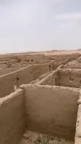 The video presents a panoramic view of an extensive archaeological site or an abandoned settlement in Saranan, Pakistan. The scene is dominated by a vast labyrinth of dilapidated mud-brick structures, most of which are reduced to low walls, suggesting former homes or buildings. The walls are made of light brown, earthen material, showing signs of age and erosion.

The camera slowly pans from left to right, revealing the sheer scale of the ruins, which stretch far into the distance under a light gray, hazy sky. The ground within and around the structures is sandy, with sparse patches of green vegetation, indicating an arid or semi-arid environment. In the background, distant hills or low mountains are visible, blending with the hazy horizon.

A few slightly more intact structures, including one with blue paint on its upper section, are visible closer to the camera, standing out subtly from the uniform brown landscape. A lone, distant tree or shrub can be seen further out in the landscape, providing the only visible greenery apart from the ground cover. The overall impression is one of an ancient or long-abandoned city or village, with a sense of historical depth and desolation.