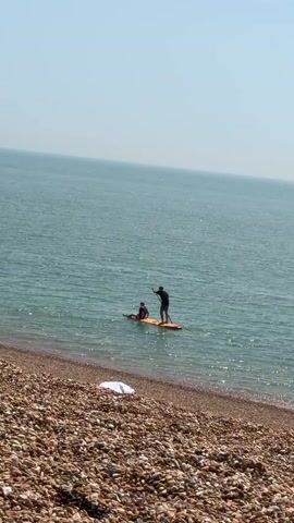 The video shows a wide shot of a pebble beach with two people on a paddle board in the ocean. The person in front on the board is seated, and the other person behind them is standing and using a paddle to move the board. The ocean is calm and the sky is clear.  A white object lies on the pebble beach in front of the camera. The scene is brightly lit. The camera is static.