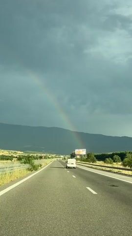 The video shows a view from a vehicle traveling on a highway. A rainbow is visible in the sky to the left, with dark, stormy clouds in the background. Mountains are visible in the distance. A white van is driving ahead on the highway. A billboard is visible on the right side of the highway. The road is asphalt and has white lane markings. The weather appears to be clearing after a storm.