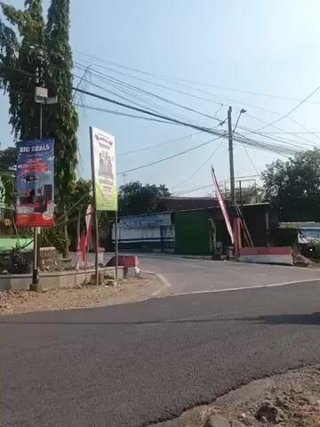 A red scooter with a rider wearing a white helmet turns onto a paved road. The rider then passes a green truck carrying a yellow road roller. The truck is driving on the right side of the road, and the scooter is in the lane to the left. The road is lined with trees and utility poles. There are also billboards on the left side of the road. The sky is clear and blue.