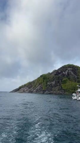 The video captures a side view from a moving boat, showing the wake behind it. In the foreground, the dark blue water is choppy. In the background, a large, white catamaran with a tall mast is anchored near a lush, green, and rocky island. The island has trees and vegetation covering its slopes. The sky above is cloudy, with some patches of lighter sky visible. The overall scene suggests a tropical, coastal environment.