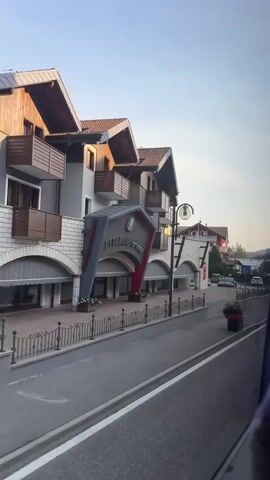 The video shows a view from a vehicle moving along a road in a European town or village. The road has white lane markings and a pedestrian crossing. On the left side of the road is a building with a prominent sign that says "BOTTAMED". The building appears to be a commercial establishment or hotel, with arched entrances and balconies. Further down the street, other buildings are visible, including a bright red building with balconies. There are several cars parked on both sides of the street and in parking lots. The background features a mountainous landscape with trees. The sky is clear and bright, suggesting daytime. The overall impression is of a quaint European setting.