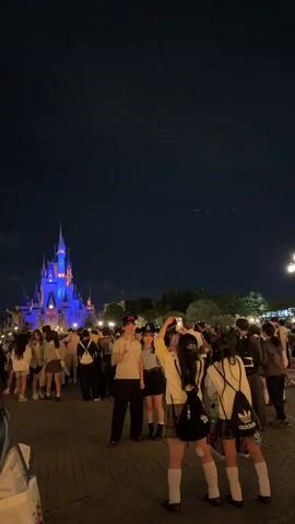 A video captured at night shows a crowd of people gathered in front of a brightly lit castle. The castle is illuminated with blue and orange lights, and its spires reach high into the dark sky. Many people in the crowd are holding up their phones, likely to take pictures or record videos. In the foreground, a group of young people are posing for a selfie. They are dressed in casual clothing, and some are wearing school uniforms. The atmosphere appears festive and lively.