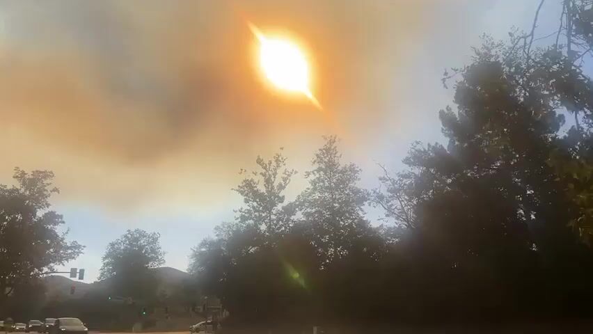 The sun shines brightly in the sky, filtered through a dense haze of smoke, likely from a distant wildfire. The smoke gives the sky a yellowish-brown cast. Trees line the lower portion of the frame. In the lower left corner, a traffic light is visible.
