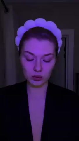 A young woman with pale skin and red-painted nails is shown from the chest up, facing the camera. She wears a white, puffy headband and a black robe. The lighting is primarily purple. She holds a pink makeup tool, examines it, and then applies it to her upper eyelid. A doorway is visible in the background, and the edge of a curtain is seen on the left side of the frame. Electronic music plays in the background.