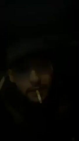 A man wearing a hat is talking to the camera. He is sitting in a dark car. He has a cigarette in his mouth. He is rapping.