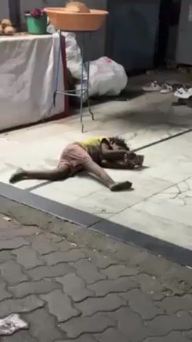 The video shows a child lying on a tiled floor in what appears to be an outdoor or semi-outdoor space. The child is wearing a yellow shirt and pink shorts, and is lying on their stomach, with their face hidden. They seem to be holding something, possibly a small box or object, with both hands. The area around the child is somewhat cluttered, with a table covered in a white cloth, a large white bag, and several pairs of shoes scattered nearby. The floor is made of light-colored tiles with dark grout lines. The lighting suggests it could be dusk or evening. The scene appears to be somewhat impoverished or unkempt.