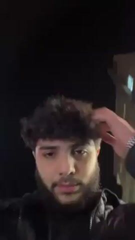 A young man with dark curly hair and a beard is seen from a low angle at night. He is wearing a dark jacket and a watch. He runs his fingers through his hair a few times and appears to be looking up at something off-camera. There is a tall building in the background. Sounds of Arabic music or conversation are audible.