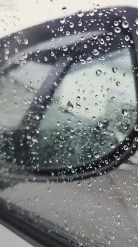 The media file shows a close-up view of a car's side mirror covered in raindrops. The mirror is reflecting a blurred, overcast sky and some faint shapes that could be trees or buildings. The raindrops are of various sizes, distorting the reflection and creating a textured surface on the glass. The overall impression is that of a rainy day. The scene is likely a street or parking area in Ballyogan, Ireland, given the context. The time of day is not clearly discernible, but the lighting suggests it could be daytime. There is no visible text in the media file. The emotion evoked is one of a typical rainy day, perhaps associated with travel or waiting.